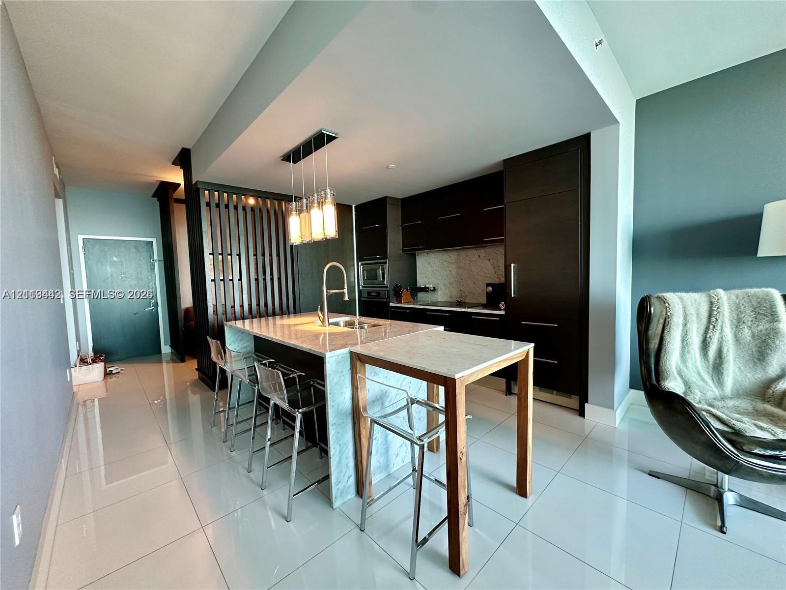 Modern kitchen featuring dark wood cabinetry, a stone-topped island with a waterfall edge, stone backsplash, built-in appliances, and a decorative vertical slat room divider