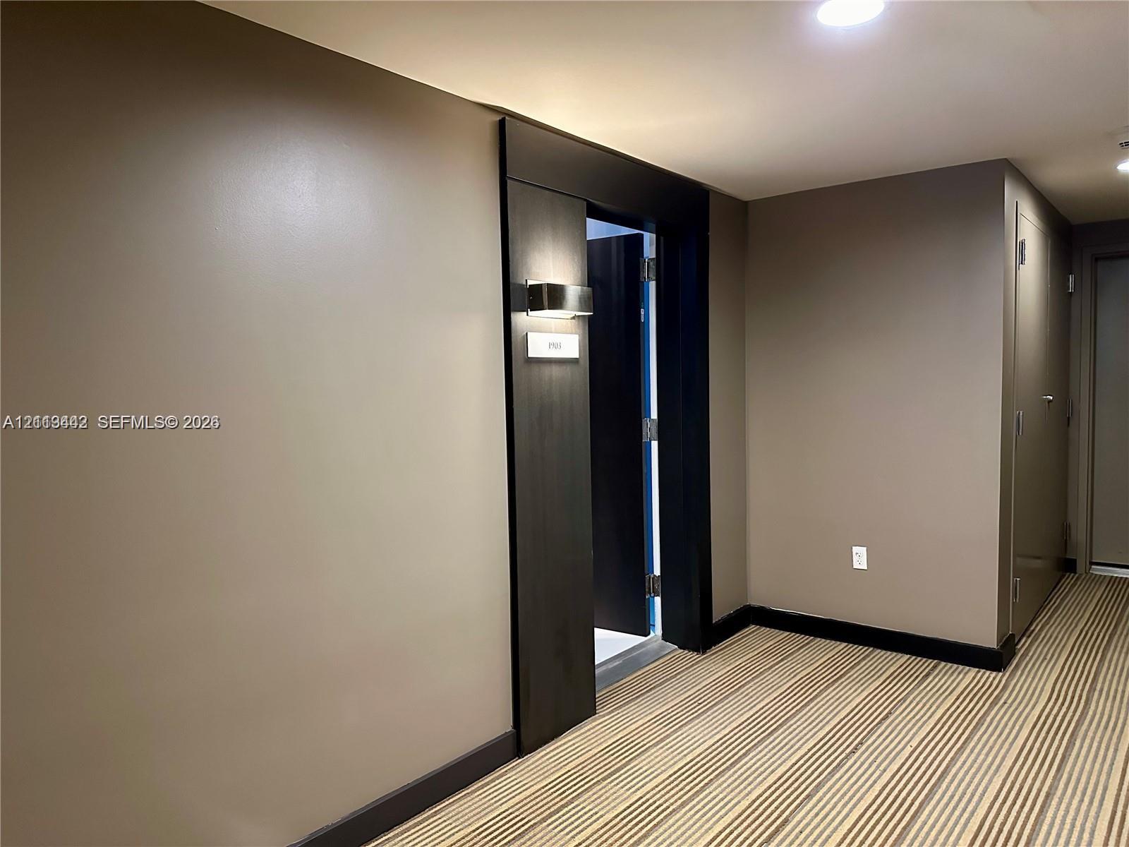 Neutral-toned interior featuring dark wood-finish trim, a recessed doorway with a wall-mounted metallic sconce, and striped carpeting