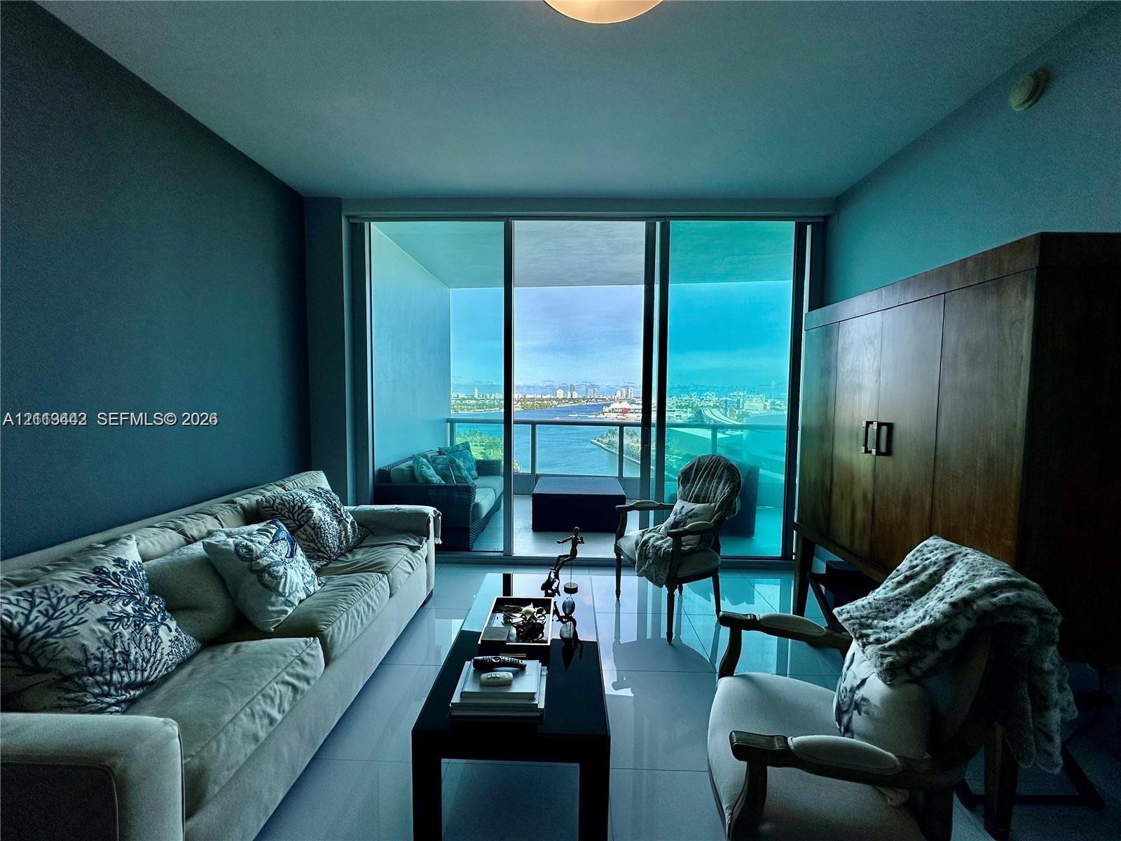 Floor-to-ceiling glass sliding doors opening to a covered balcony, large-format tile flooring, and panoramic water scenery