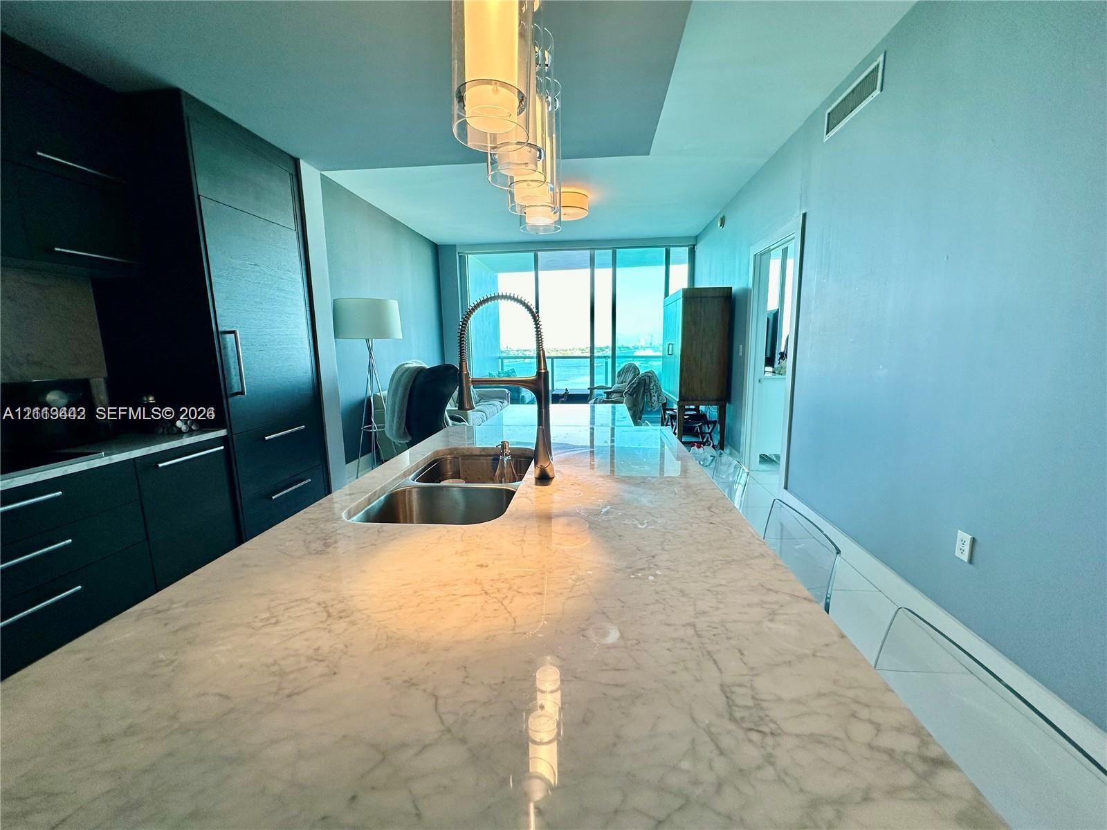 Kitchen featuring a stone-surface island, undermount stainless steel sink, pull-down chrome faucet, dark cabinetry, and glass pendant lighting