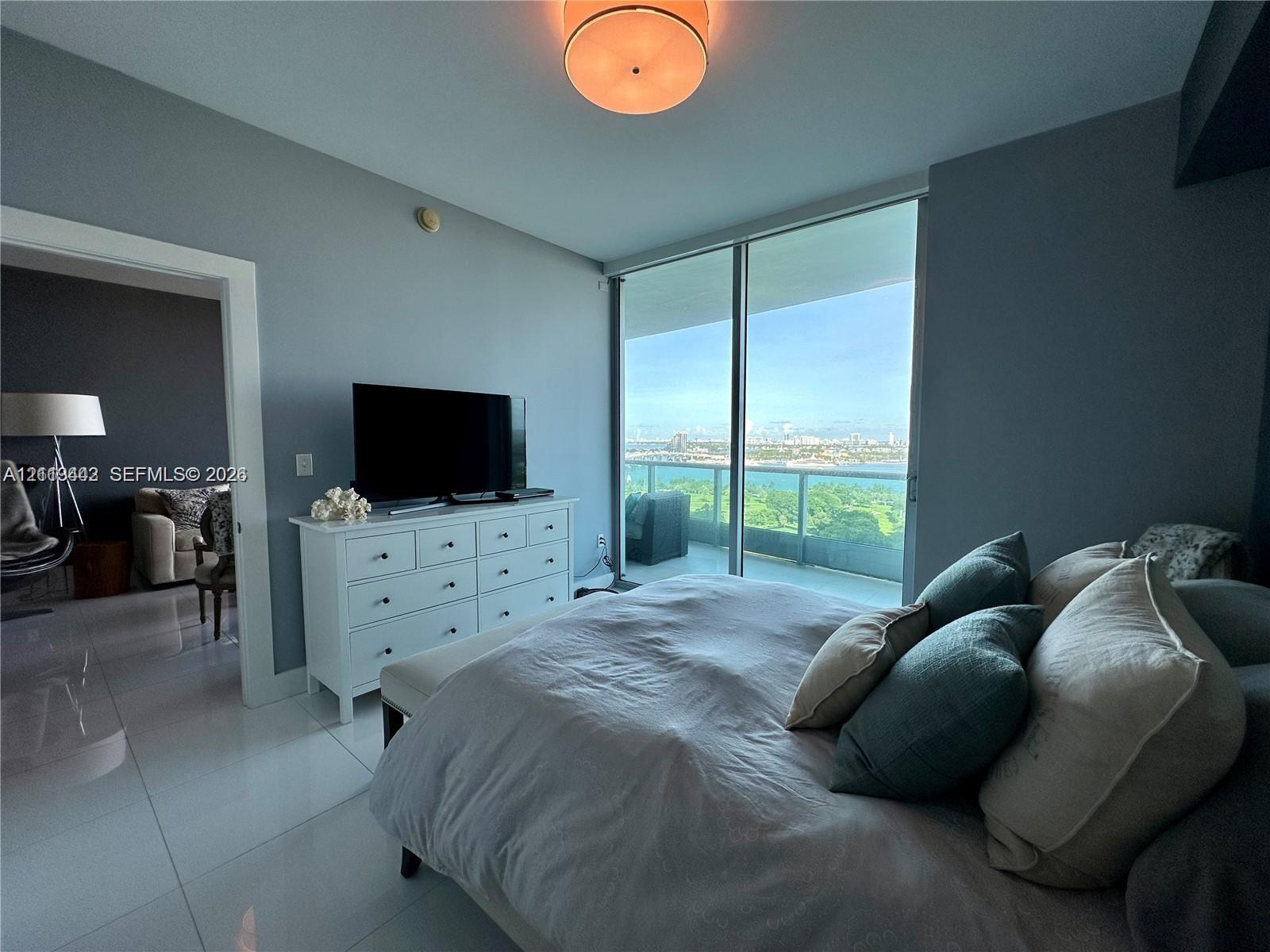 Floor-to-ceiling glass sliding doors, private balcony, neutral large-format tile flooring, and a circular flush-mount ceiling light fixture