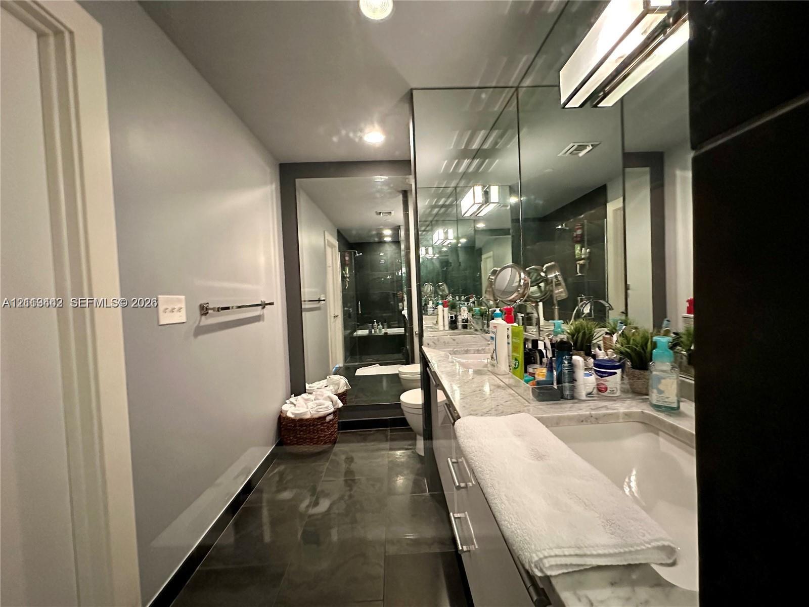 Stone-topped vanity with an integrated rectangular sink, large full-width wall mirror, dark tile flooring, and integrated linear lighting