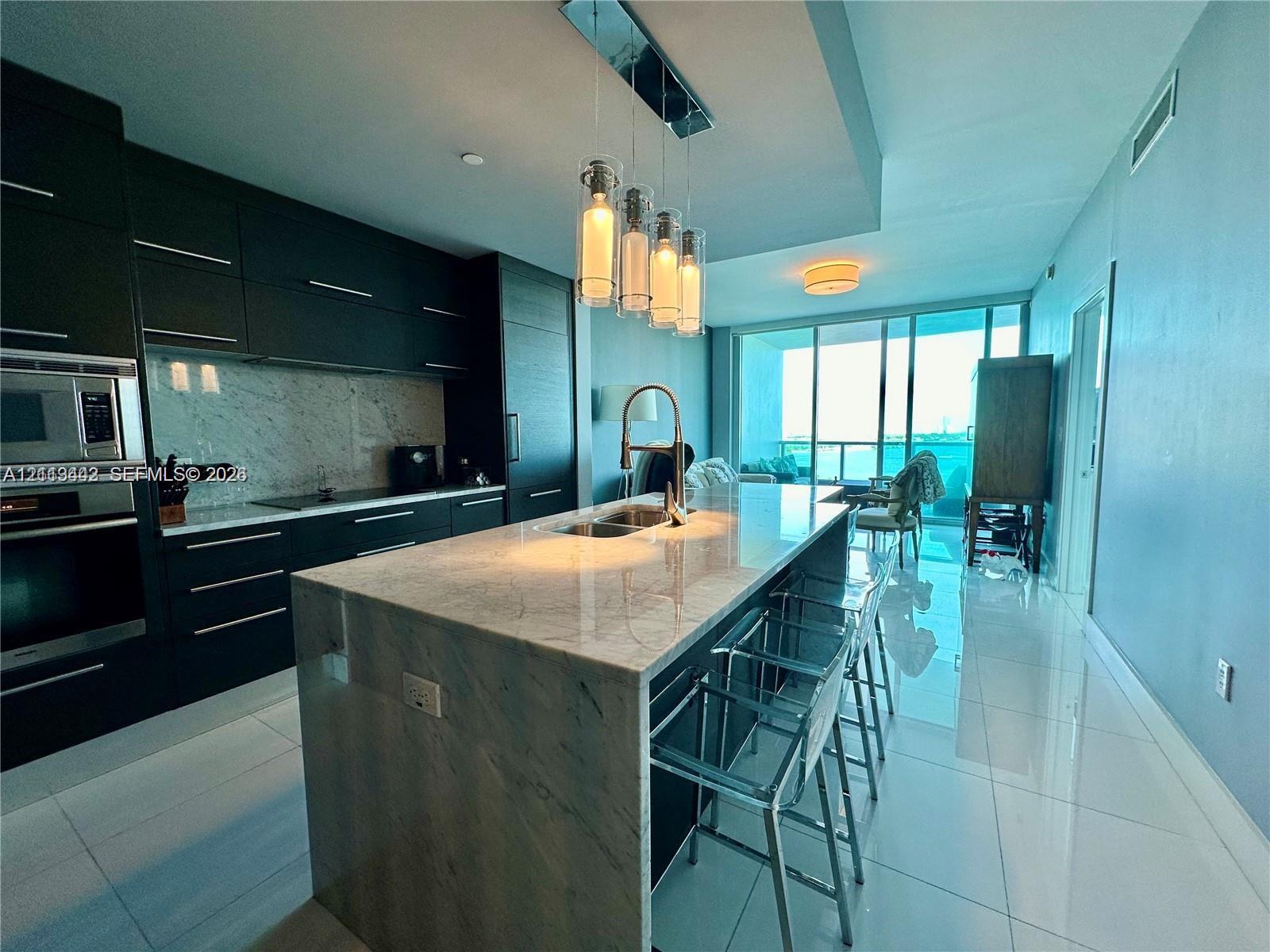 Modern kitchen featuring matte black flat-panel cabinetry, stone countertops with waterfall edge, integrated stainless steel appliances, multi-pendant island lighting, and large-format polished tile flooring