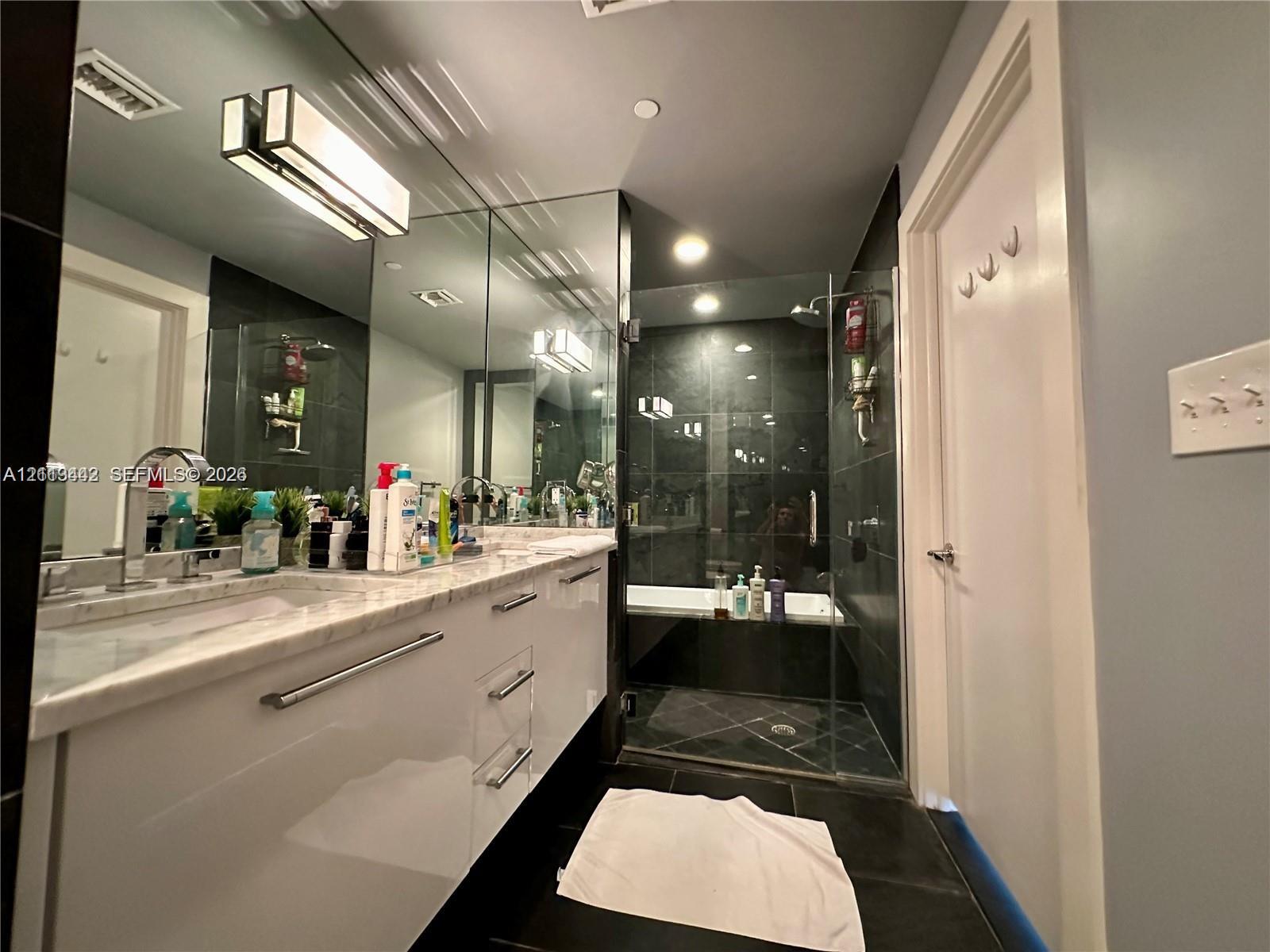 Glossy white cabinetry with metal hardware, stone-finish countertop, full-wall mirror, and glass-enclosed shower with dark tile surround