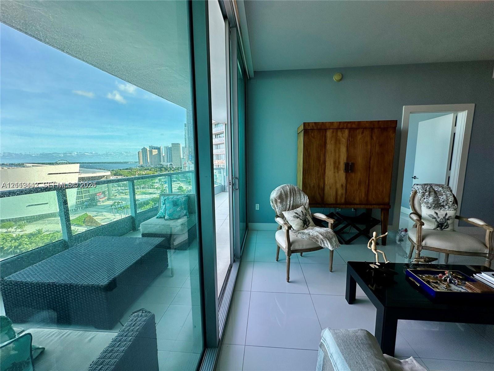 Floor-to-ceiling glass sliding doors opening to an expansive balcony with glass railings, large-format tile flooring, and coastal skyline scenery