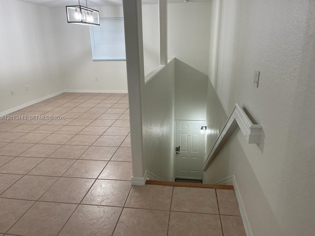 Ceramic tile flooring throughout the open interior space, featuring a modern rectangular pendant light fixture and a stairway with white wood handrails leading to an exterior entrance door
