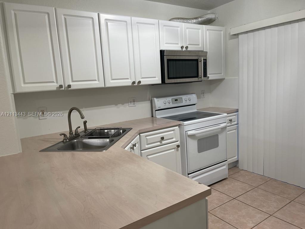 Kitchen featuring white shaker-style cabinetry, a stainless steel over-the-range microwave, a neutral-toned countertop, a double-basin stainless steel sink with a high-arch faucet, and ceramic tile flooring Kitchen featuring white shaker-style cabinetry, a stainless steel over-the-range microwave, a neutral-toned countertop, a double-basin stainless steel sink with a high-arch faucet, and ceramic tile flooring