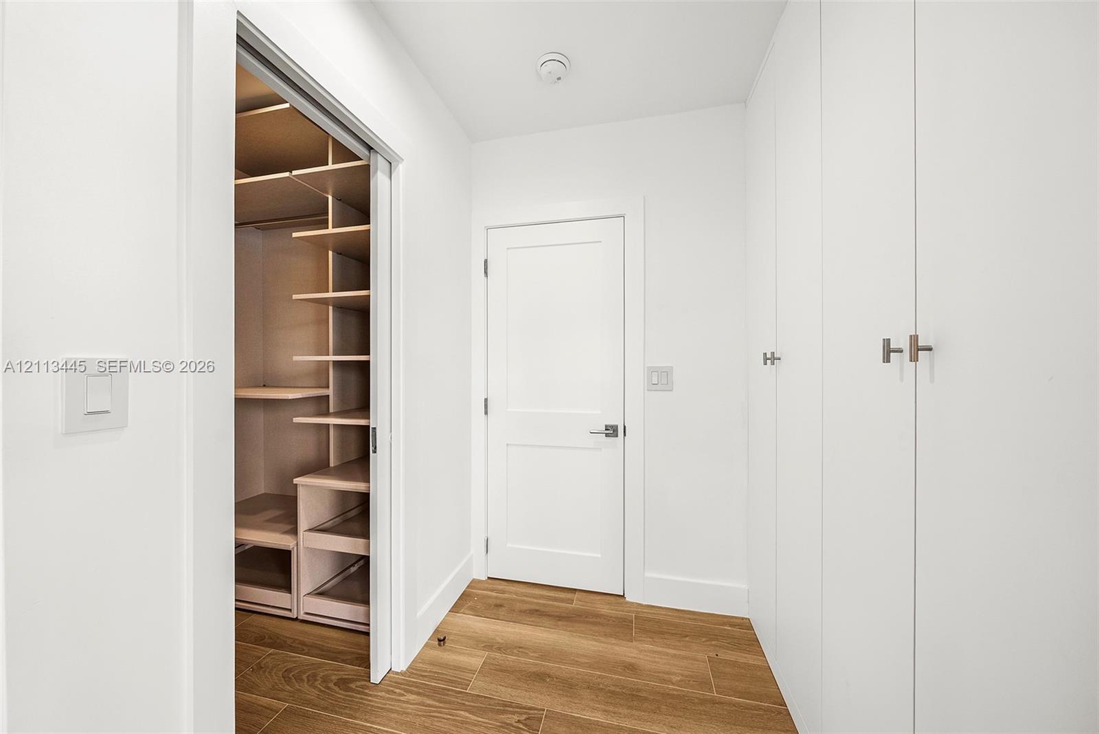 Walk-in closet featuring custom shelving and pull-out drawers, complemented by floor-to-ceiling built-in cabinetry, white shaker-style doors, and wood-finish flooring
