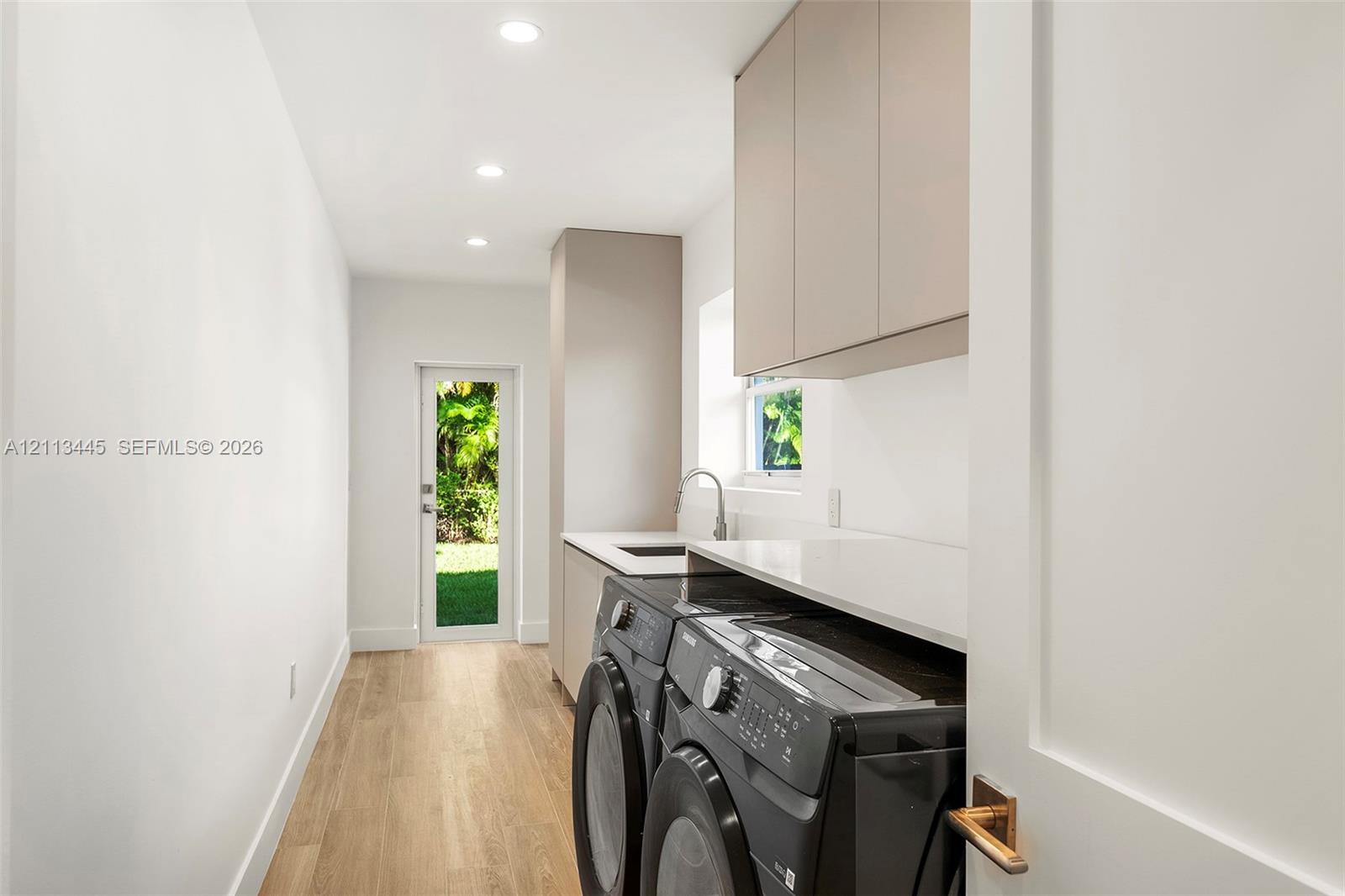 Laundry area featuring minimalist flat-panel cabinetry, light-toned stone-finish countertops, an undermount sink with a gooseneck faucet, wood-finish flooring, and recessed ceiling lighting
