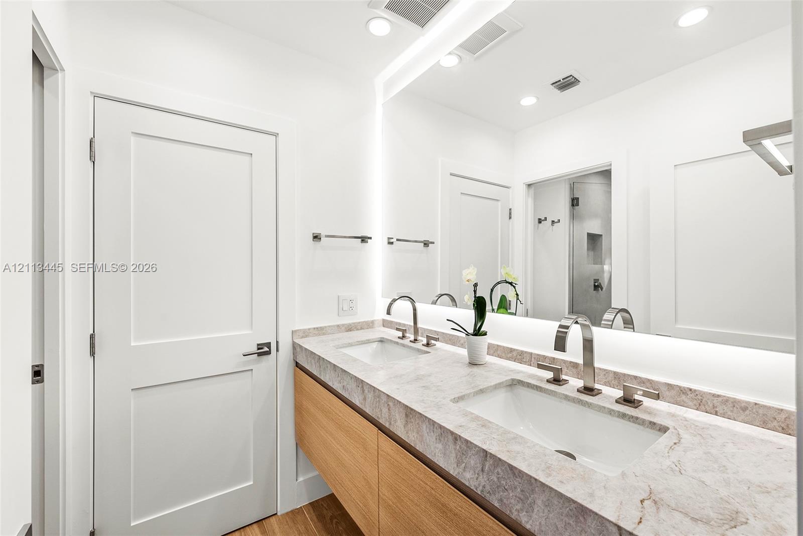 Floating vanity with wood-finish cabinetry, stone countertop, integrated dual undermount sinks, tall arch-neck faucets, and large wall-to-wall mirror with integrated lighting