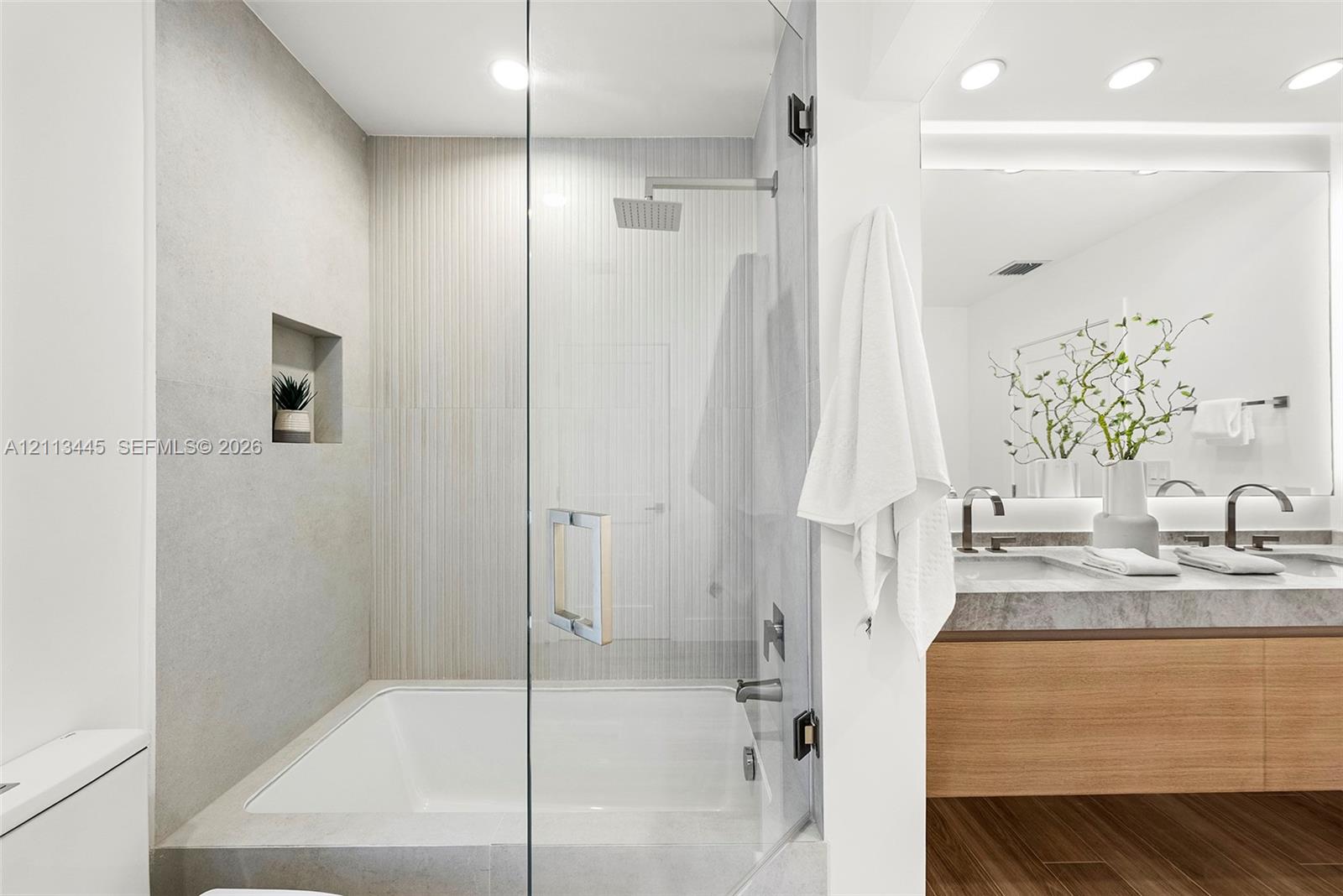 Shower and tub combination featuring glass enclosure, rain-style shower head, built-in recessed wall niche, dual-sink vanity with stone-finish countertop, and wood-finish flooring