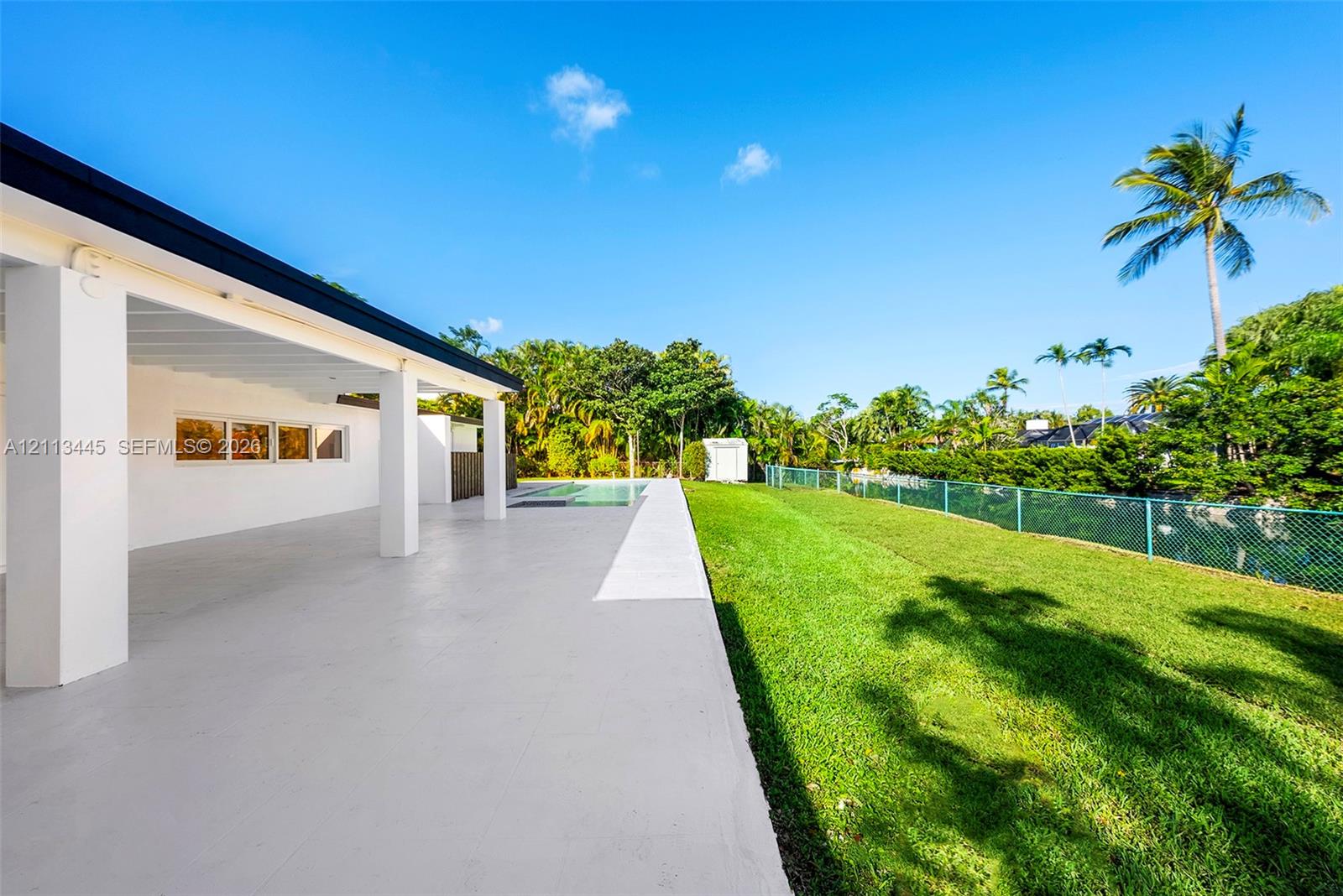 Covered concrete patio featuring white support columns, inground pool, manicured lawn, and perimeter fencing