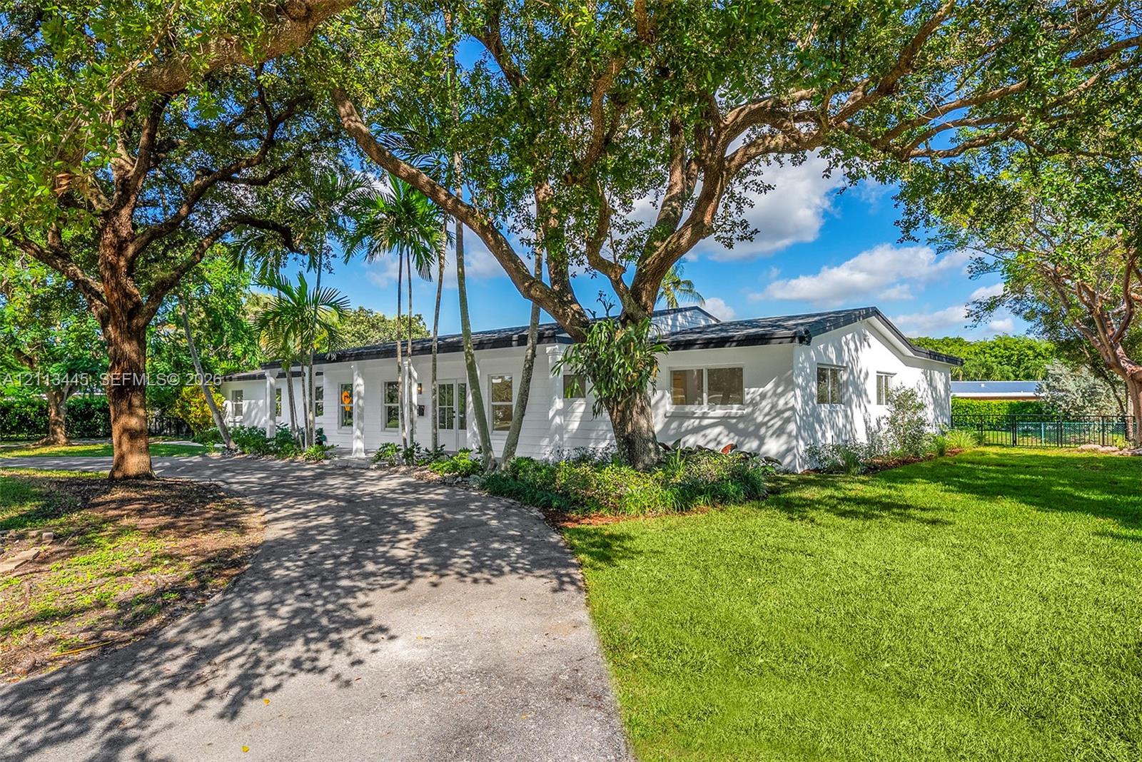 Single-story exterior featuring a white masonry faade, a paved circular driveway, mature oak trees, and palm landscaping