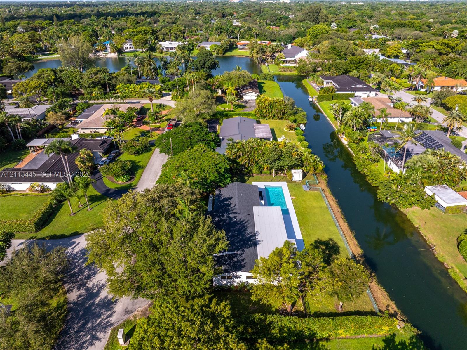 Aerial perspective of a waterfront property featuring a swimming pool, expansive lawn, mature tree canopy, and canal frontage