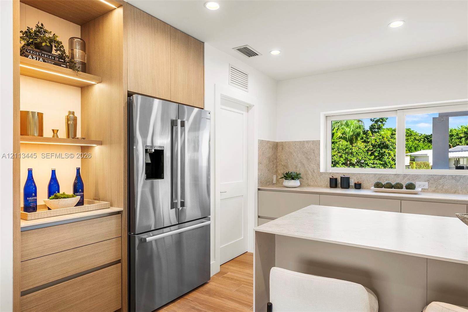 Kitchen featuring flat-panel cabinetry, light-toned stone-finish countertops, stainless steel refrigerator, integrated wood-finish shelving with recessed lighting, and large horizontal window