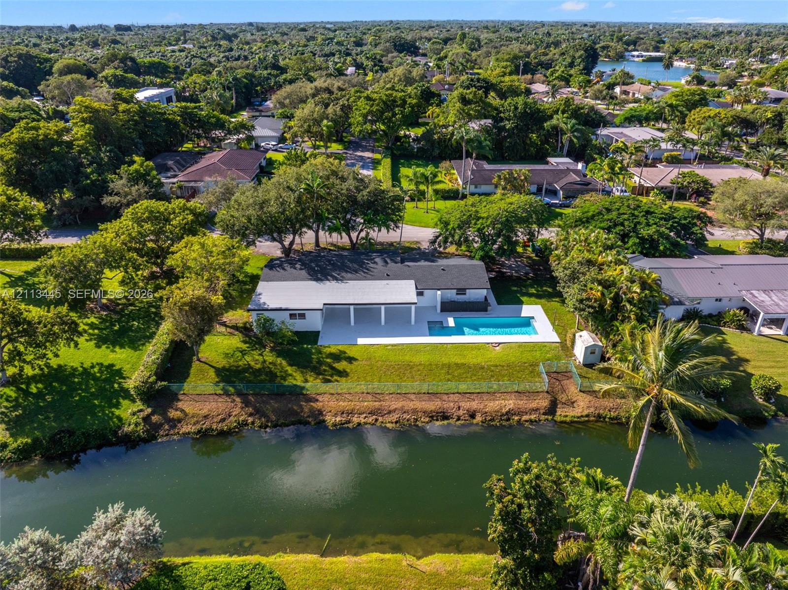 Waterfront residence featuring a rectangular swimming pool, covered patio, sprawling lawn, and mature trees
