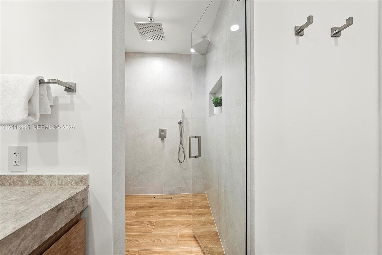 Walk-in shower featuring glass enclosure, ceiling-mounted rainfall showerhead, handheld sprayer, integrated wall niche, and wood-finish flooring