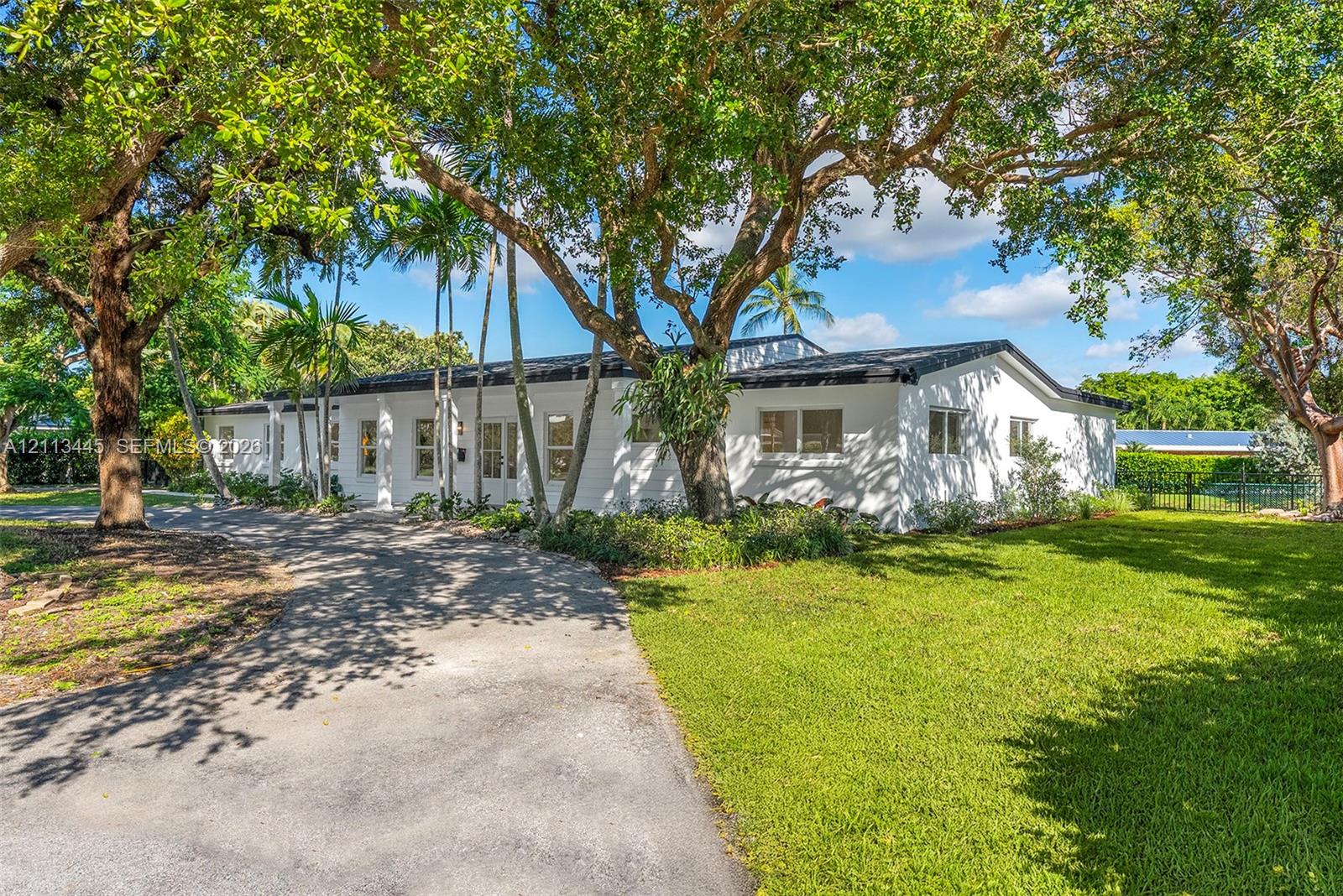Single-story exterior featuring white masonry walls, a flat roofline, paved driveway, and mature landscaping