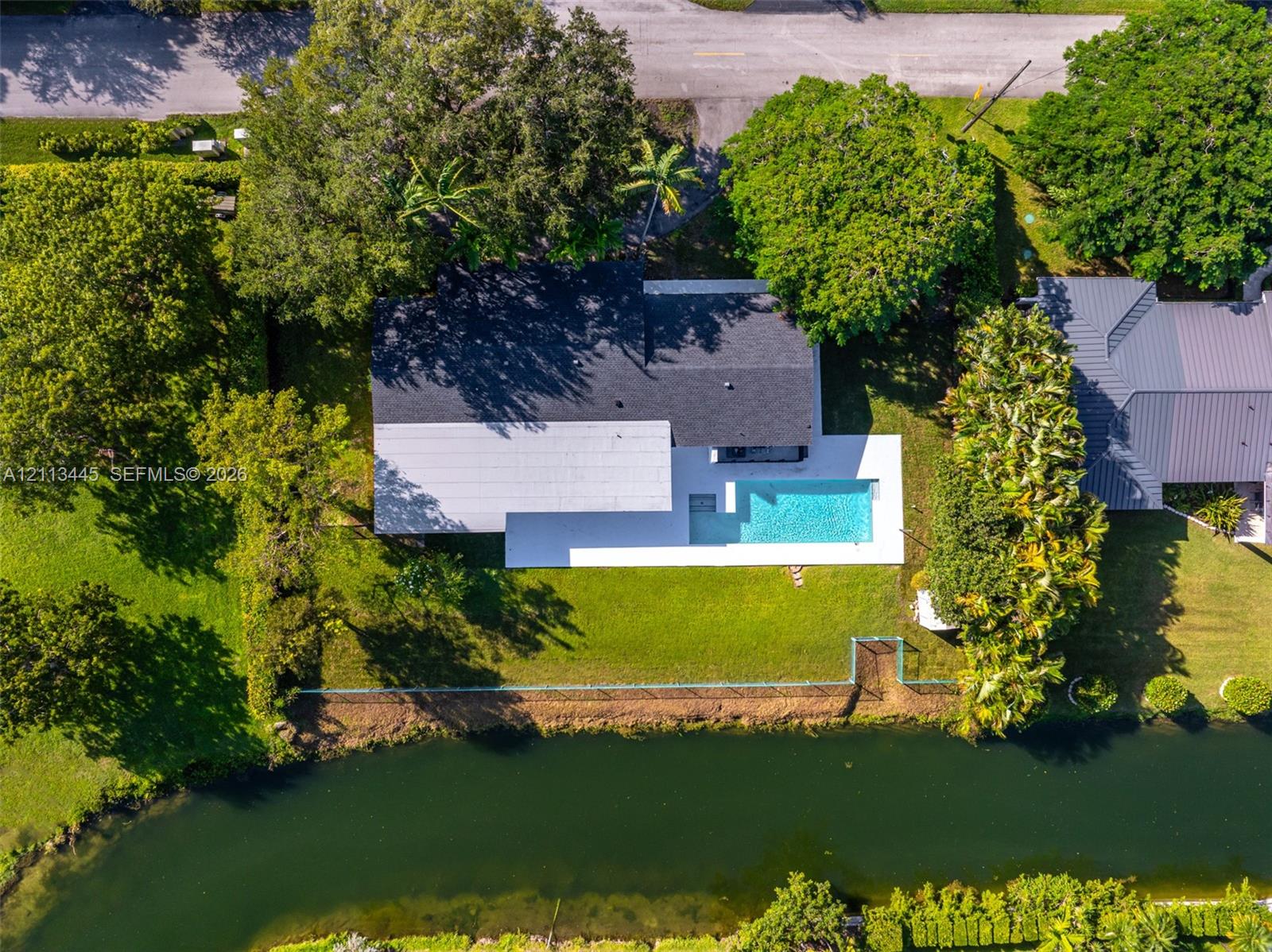 Aerial perspective featuring a waterfront property with an in-ground swimming pool, concrete pool deck, expansive lawn, and mature landscaping