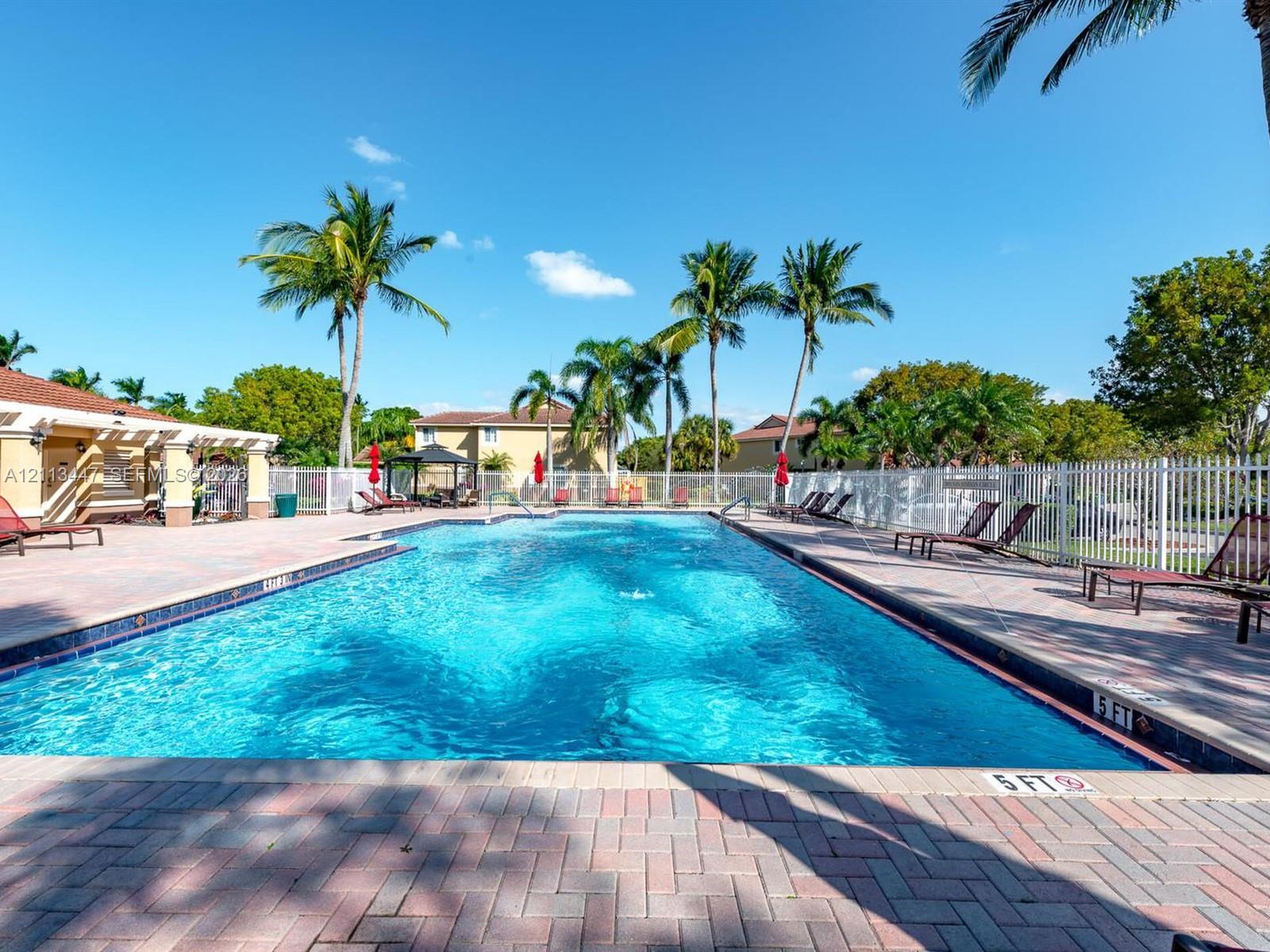 Rectangular swimming pool with brick paver decking, surrounded by white perimeter fencing and mature palm trees