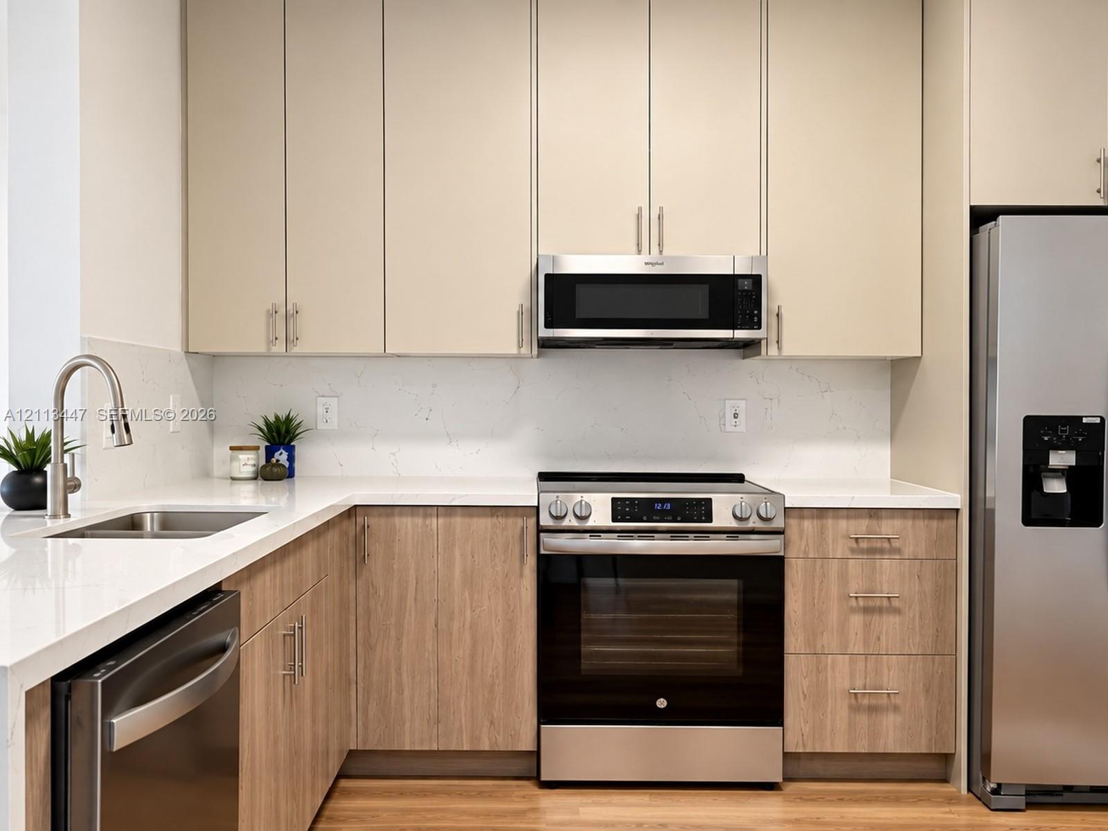 Kitchen featuring flat-panel, luxury laminated flooring, base cabinetry, light-toned upper cabinetry, Carrara quartz countertops with waterfall edge, full-height solid-surface backsplash, and stainless steel appliances