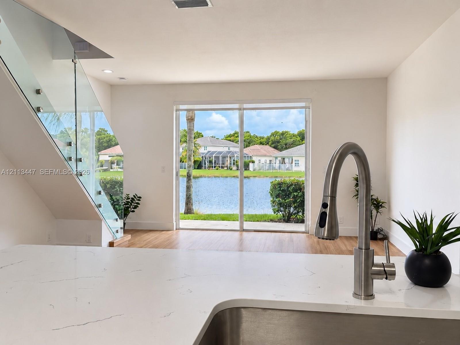 Kitchen featuring white Carrara quartz countertops, stainless steel undermount sink with arched pull-down faucet, glass-panel staircase railing, and sliding glass doors leading to water-side exterior
