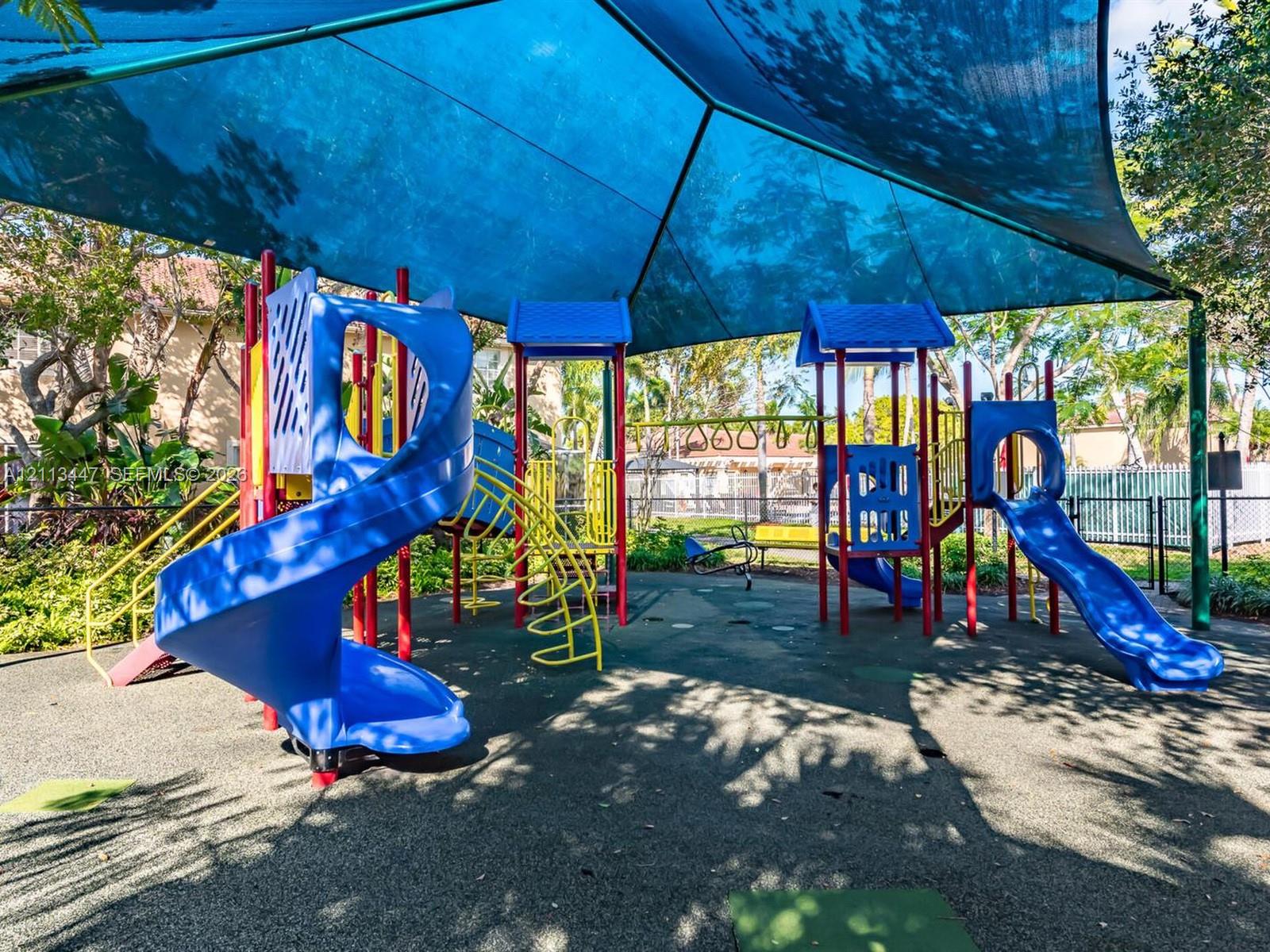 Playground area featuring a multi-level structure with spiral and straight slides, overhead ladder, protective shade canopy, and poured-in-place rubber surfacing