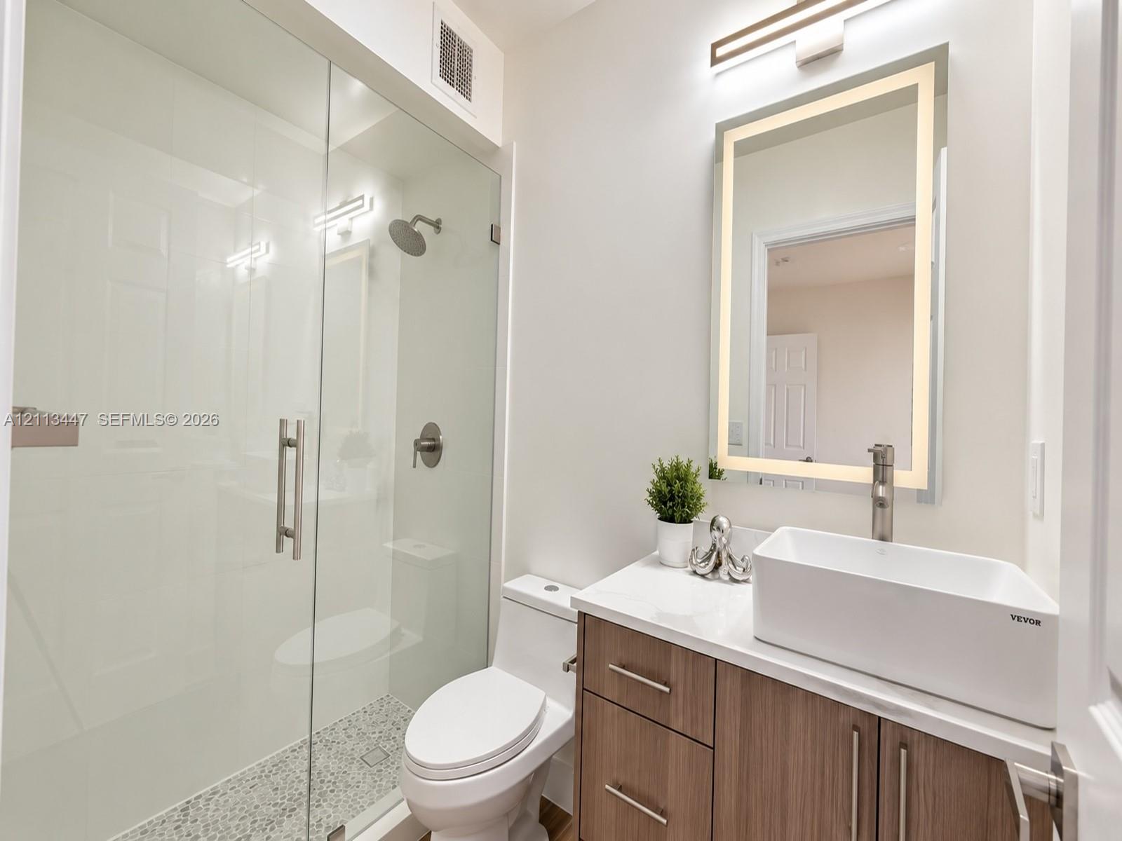 Bathroom featuring a glass-enclosed shower with pebble-tile flooring, a wood-finish vanity with a vessel sink, and an integrated LED-lighted mirror