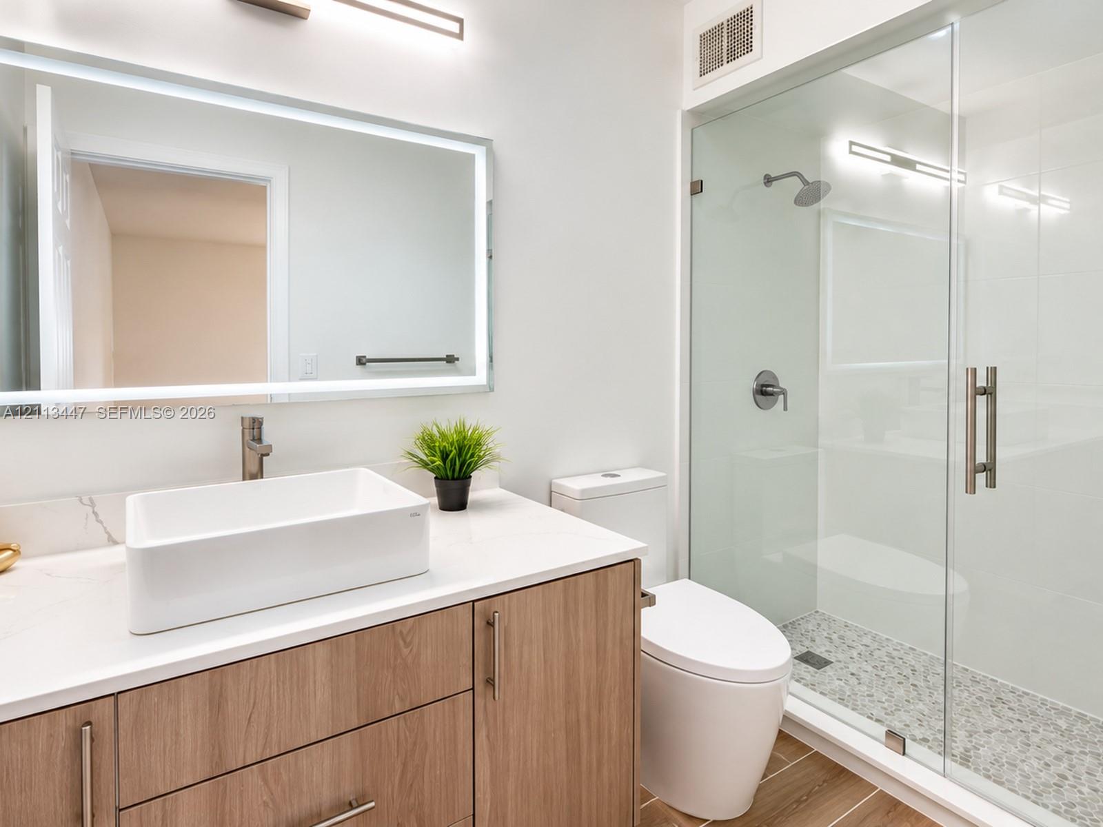 Bathroom featuring a wood-finish vanity with a stone-top counter, white vessel sink, rectangular LED-backlit mirror, and a walk-in shower with a glass enclosure and stone tile flooring
