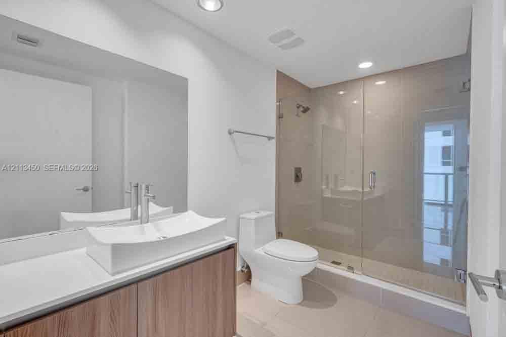 Bathroom featuring a wood-finish floating vanity with a white vessel sink, large wall-mounted mirror, recessed lighting, and a glass-enclosed walk-in shower with tile flooring