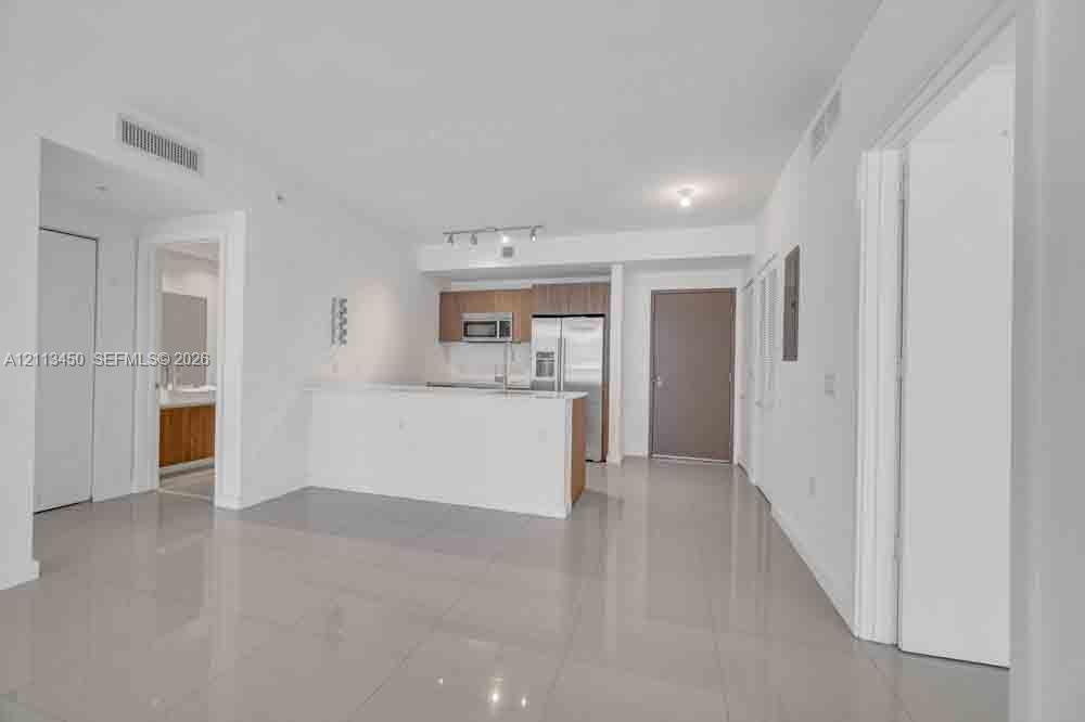 Open-concept interior featuring light-toned tile flooring, kitchen with stainless steel appliances, wood-finish cabinetry, and a breakfast bar