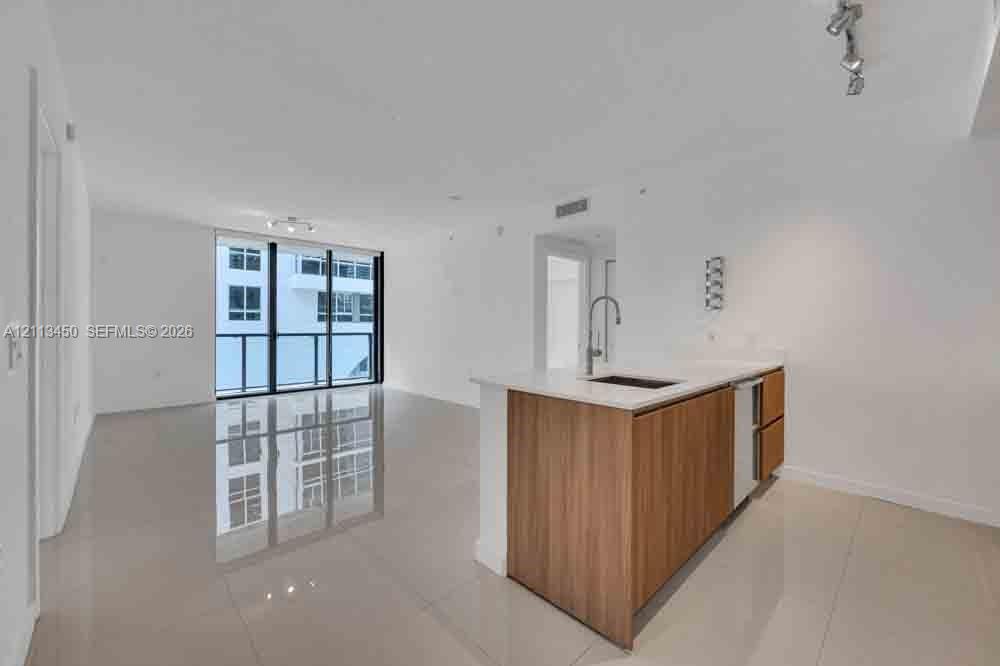 Open-concept interior featuring large-format tile flooring, wood-finish kitchen cabinetry, a white countertop with an integrated sink, and floor-to-ceiling glass sliding doors