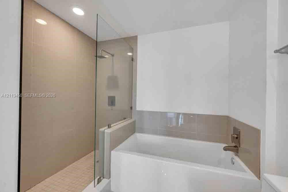 Walk-in shower with large-format wall tile, frameless glass enclosure, and ceiling-mounted rain showerhead, paired with a deep soaking tub featuring metallic fixtures and tile surround