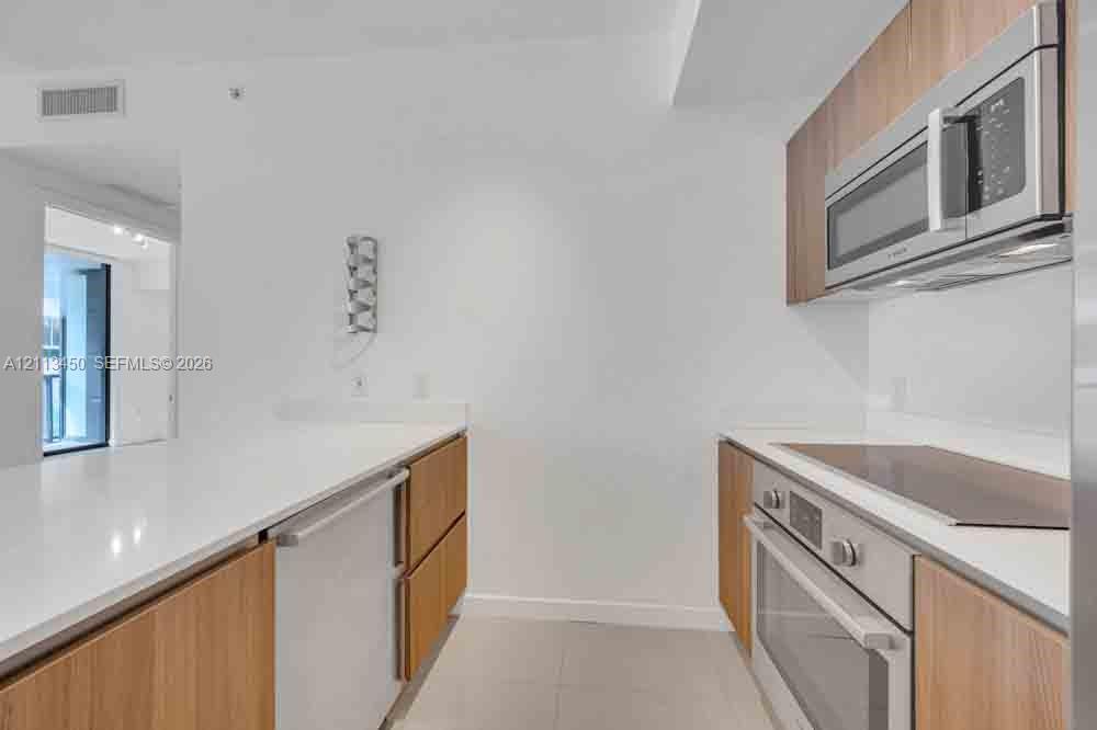 Kitchen featuring wood-finish cabinetry, white solid-surface countertops, a stainless steel over-the-range microwave, a built-in oven, and a flat-surface cooktop