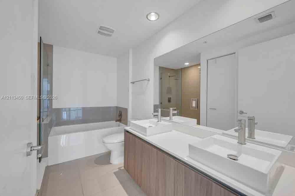 Double vanity featuring wood-finish cabinetry, white solid-surface countertops, and rectangular vessel sinks with polished chrome faucets, complemented by a large wall-spanning mirror and a deep soaking tub with neutral tile surround