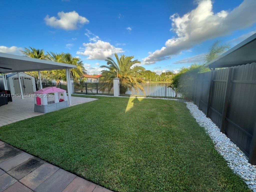 Rear yard featuring a covered patio, paved hardscaping, manicured lawn, dark metal fencing, and stone landscape bordering