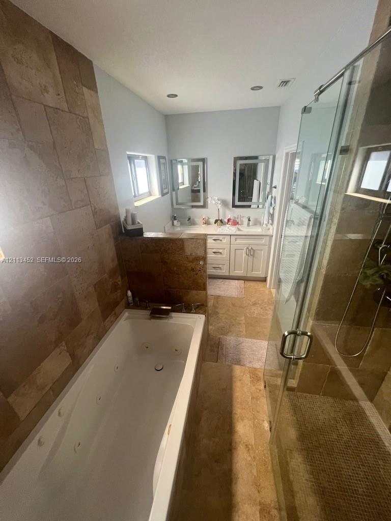 Jetted soaking tub with tile surround, walk-in glass-enclosed shower, dual-sink vanity with cabinetry, and stone-finish flooring