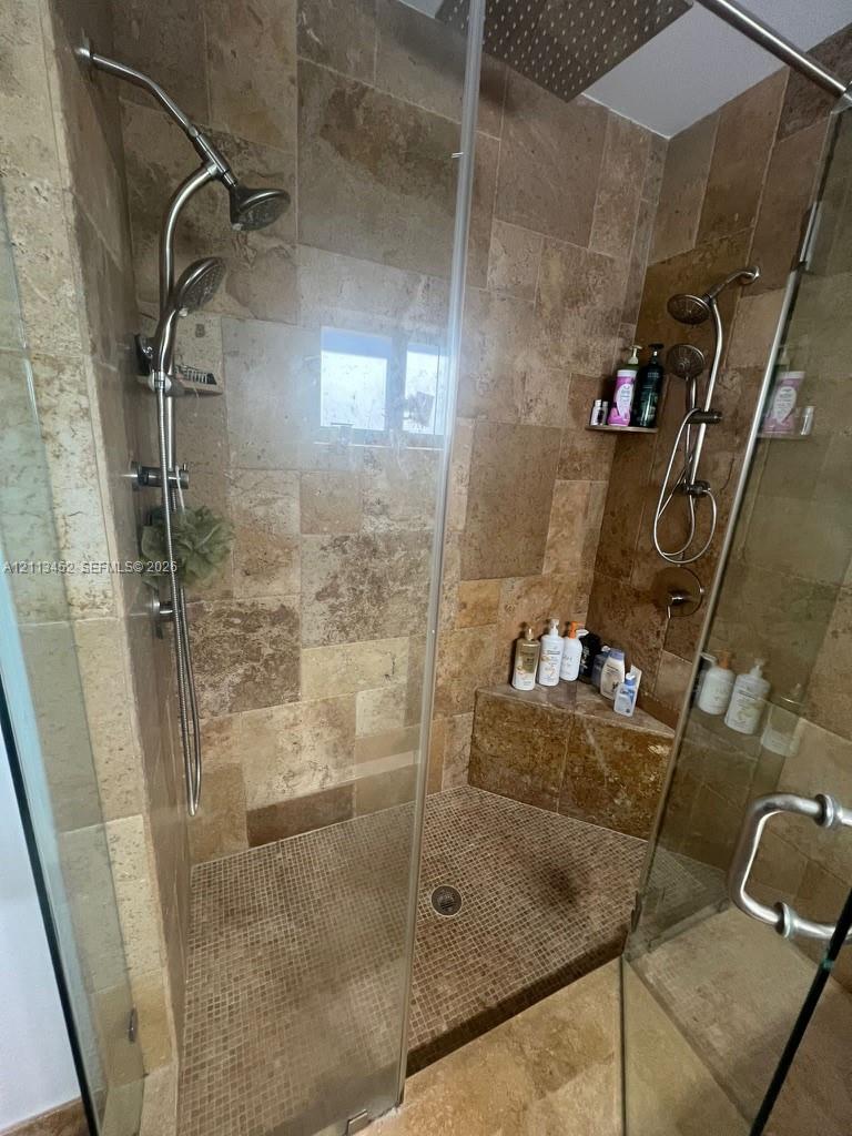 Walk-in shower featuring natural stone wall tile, mosaic tile flooring, built-in corner bench, dual adjustable handheld showerheads, and frameless glass enclosure