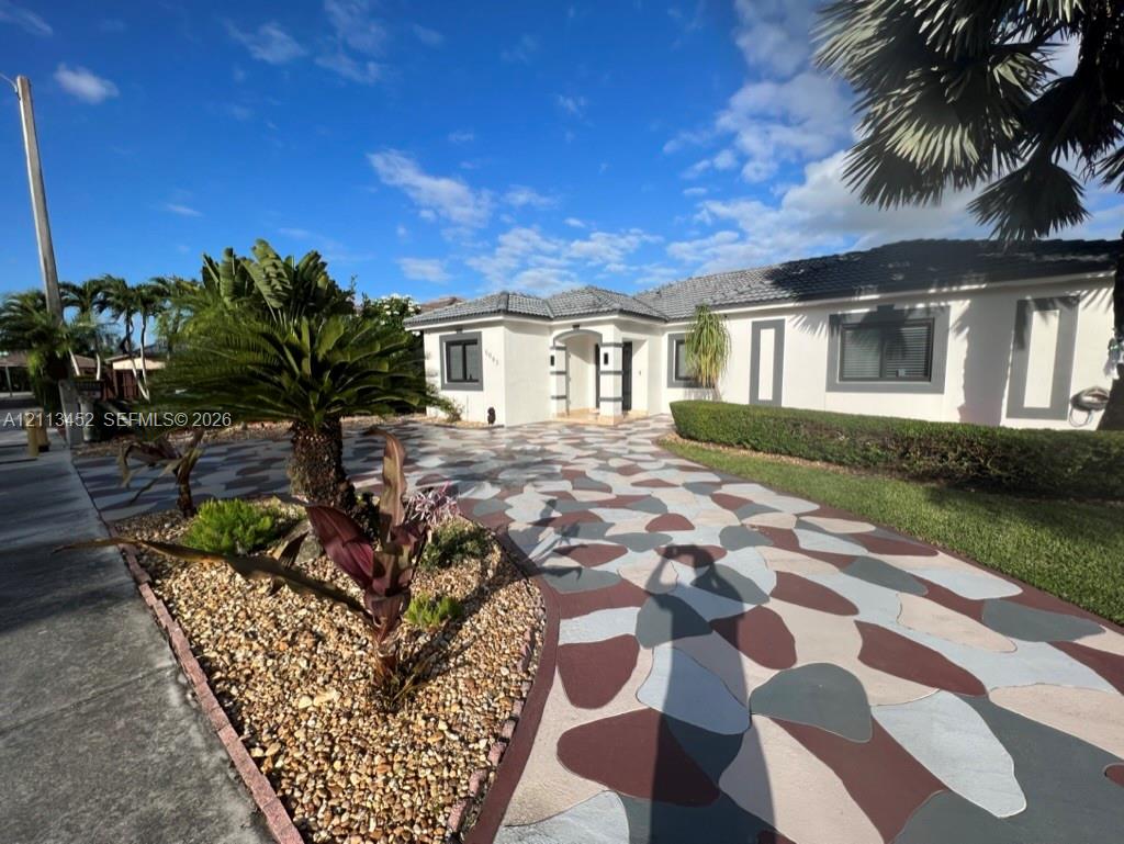 Single-story exterior featuring a decorative stone-pattern driveway, concrete facade, hipped tile roof, and landscaped garden beds with established tropical flora