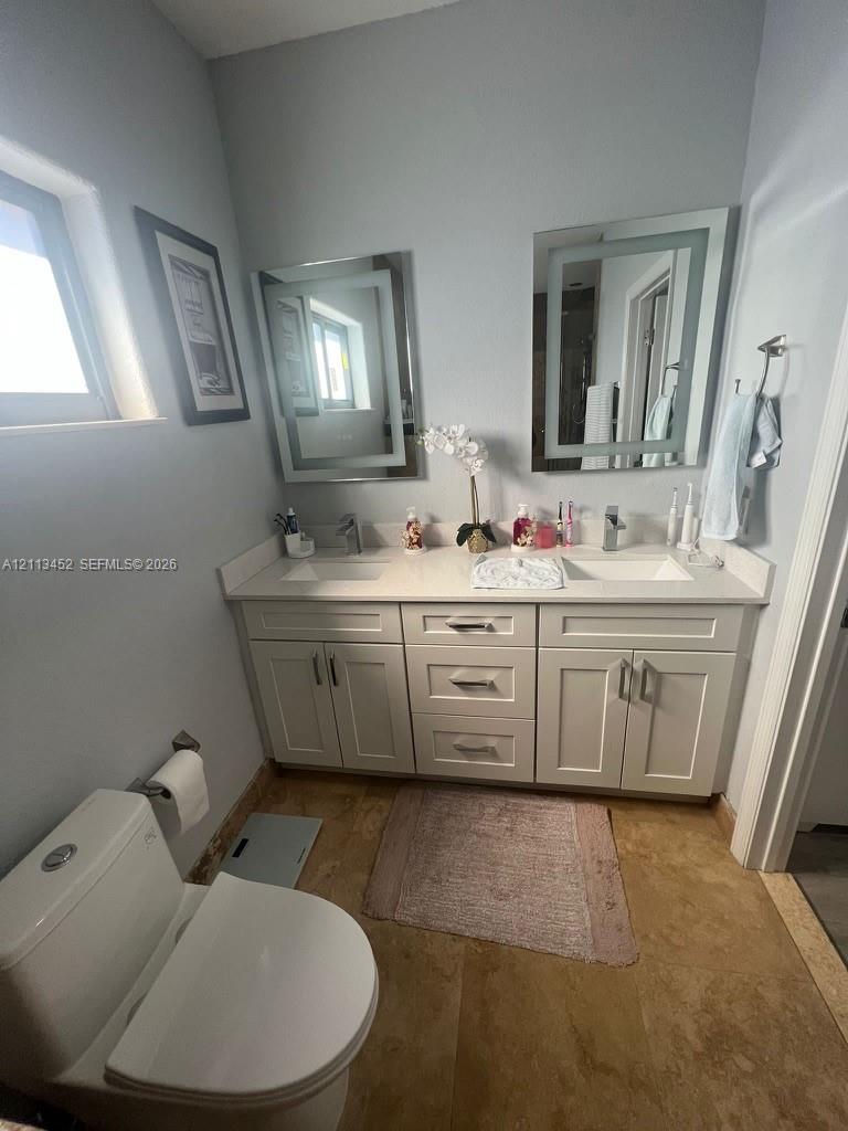 Dual-sink vanity with white shaker-style cabinetry, solid-surface countertop, integrated rectangular basins, and dual lighted wall-mounted mirrors, complemented by stone-finish flooring