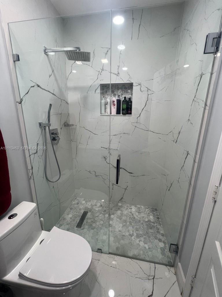 Walk-in shower featuring large-format veined tile surround, frameless glass enclosure, rainfall showerhead with handheld attachment, recessed tile niche, and textured pebble floor tiling with a linear drain
