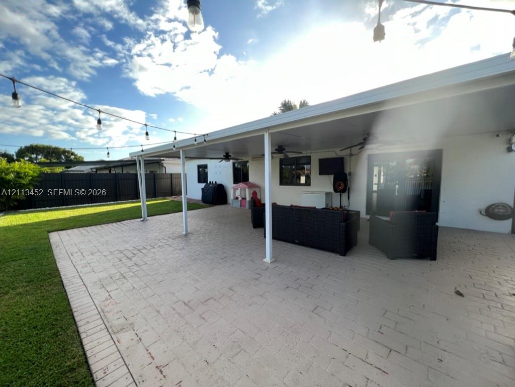 Covered outdoor patio featuring stamped concrete flooring, integrated ceiling fans, overhead string lighting, and a manicured lawn enclosed by perimeter fencing