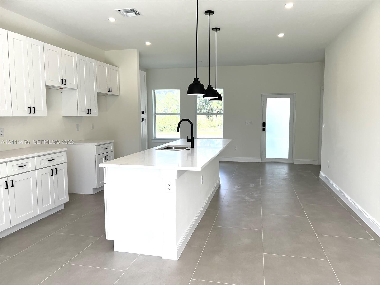 Kitchen featuring white cabinetry, an expansive center island with a stone-surface countertop, matte black faucet hardware, pendant lighting, and neutral-toned tile flooring Kitchen featuring white cabinetry, an expansive center island with a stone-surface countertop, matte black faucet hardware, pendant lighting, and neutral-toned tile flooring