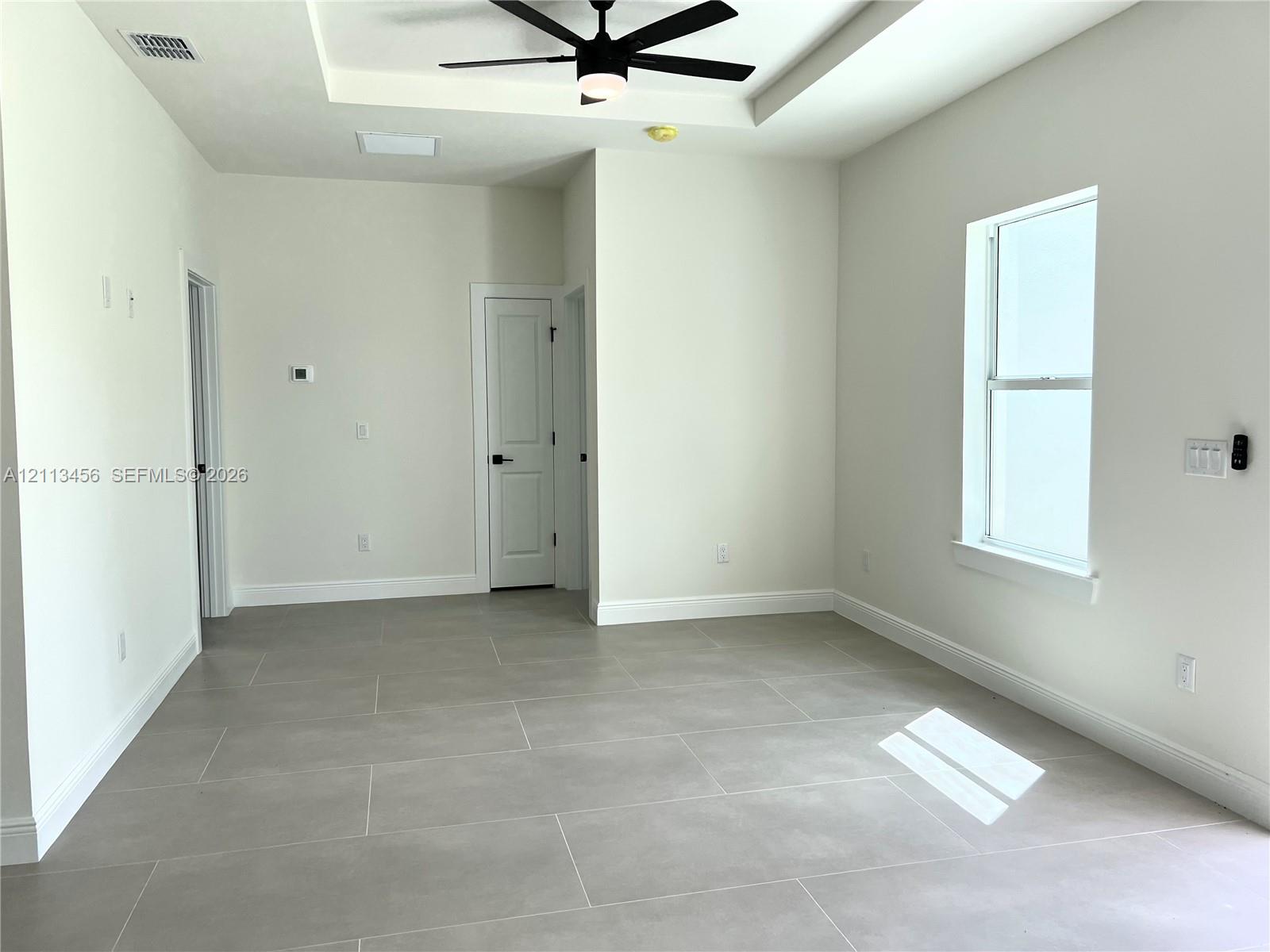 Tray ceiling featuring a central matte black ceiling fan, complemented by large-format neutral tile flooring and a vertical window Tray ceiling featuring a central matte black ceiling fan, complemented by large-format neutral tile flooring and a vertical window