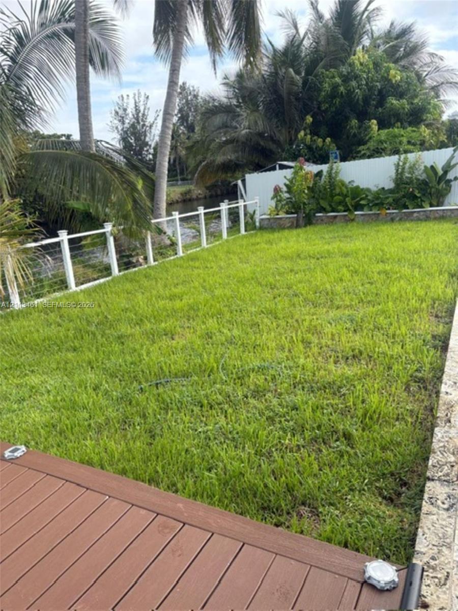 Backyard featuring a wood-finish deck, natural grass lawn, white post-and-cable perimeter fencing, stone retaining wall, and mature palm trees