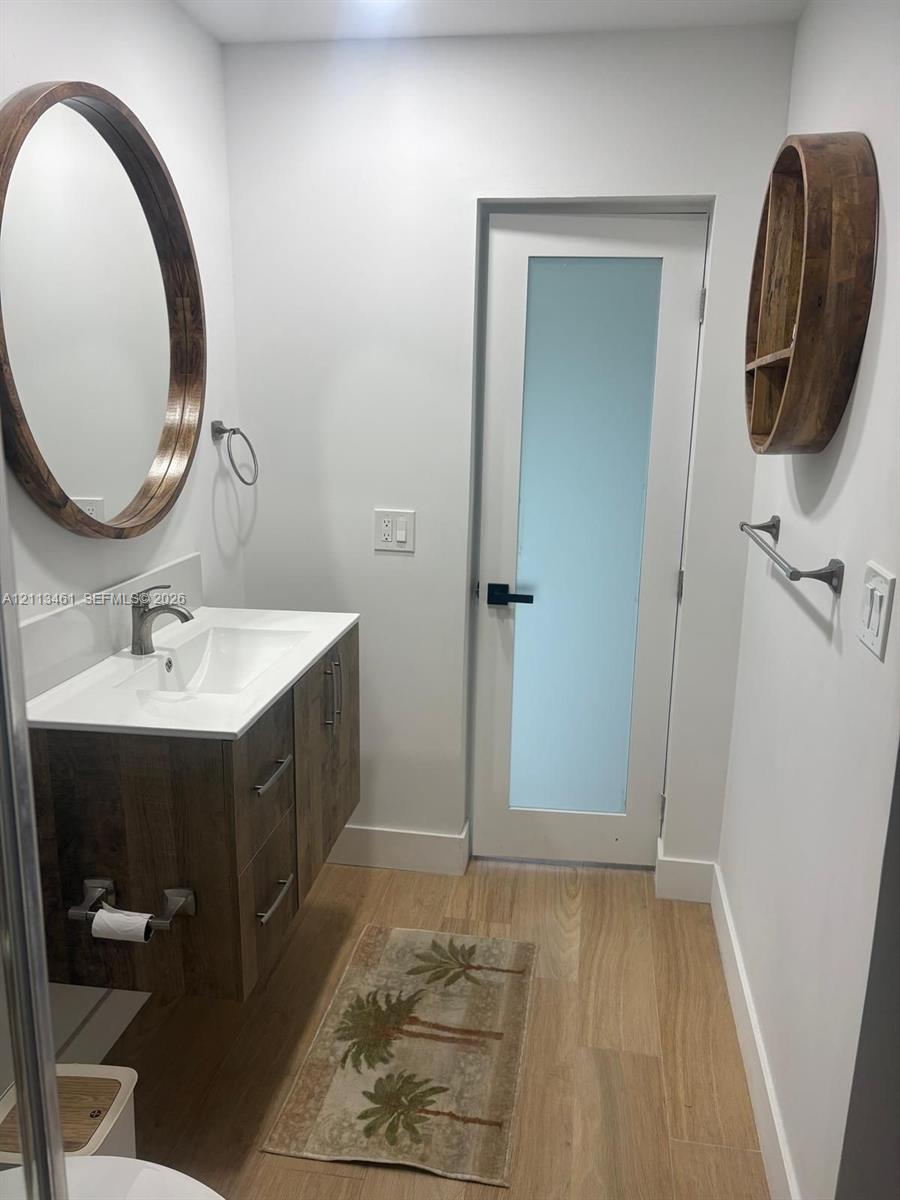 Floating wood-finish vanity with an integrated basin, round wood-frame mirror, wood-finish wall shelving, wood-finish flooring, and a single-panel door with a frosted glass insert