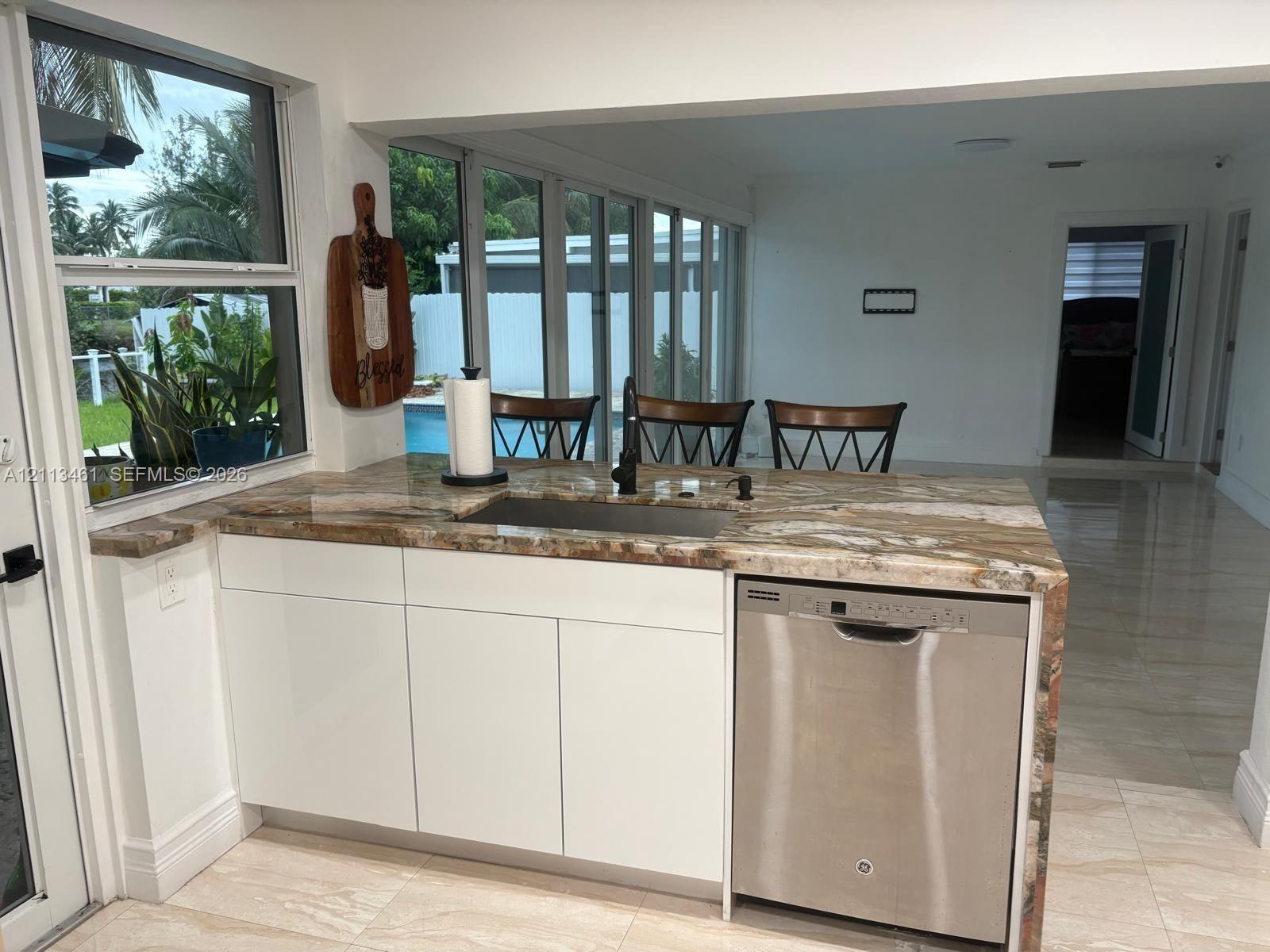 Kitchen island featuring natural stone waterfall countertops, flat-panel white cabinetry, a stainless steel dishwasher, and an under-mount sink with a matte black faucet