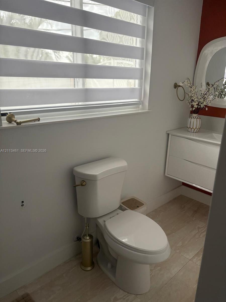 Floating white vanity with integrated basin, light-toned tiled flooring, adjustable horizontal window shade, and brushed metallic hardware
