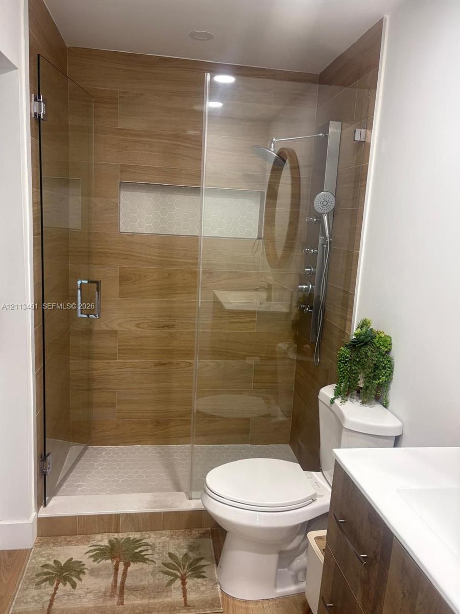 Walk-in shower featuring wood-finish tile surround, integrated mosaic niche, frameless glass enclosure, and multi-function shower system with handheld wand, paired with a wood-finish vanity and white countertop