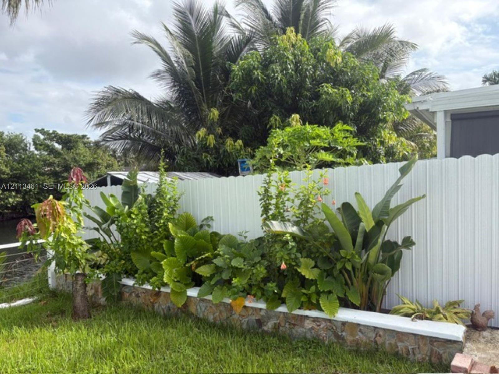 Raised stone-veneer garden bed with a white cap, accented by mature tropical landscaping and a white vinyl privacy fence